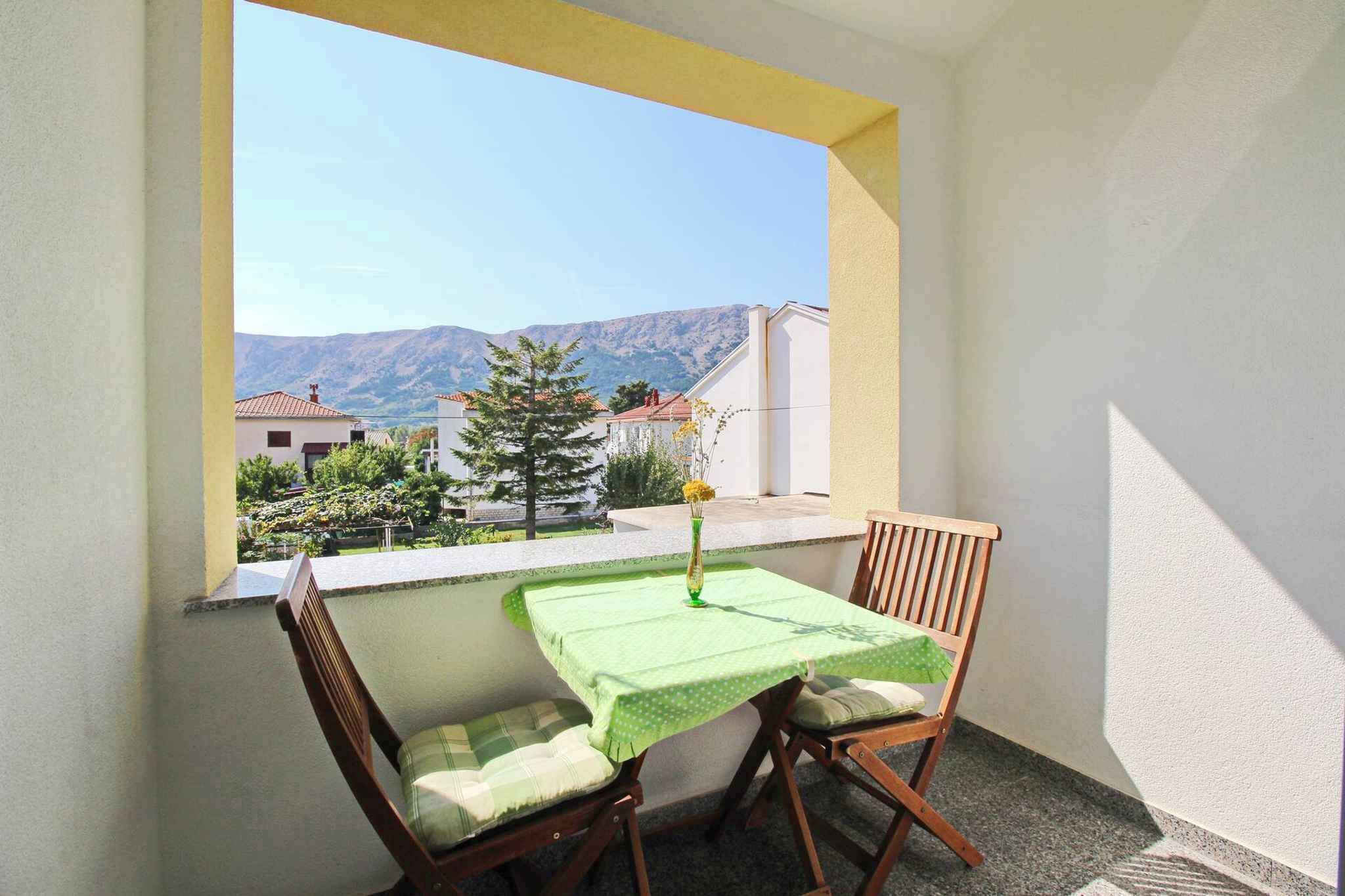 Varanda com mesa e duas cadeiras, um vaso com flores e vista para casas e montanhas.