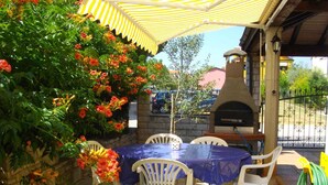 Eine Terrasse im Freien mit Tisch und Stühlen unter einem gestreiften Markisenstoff, umgeben von blühenden Pflanzen und einem Steinofen.