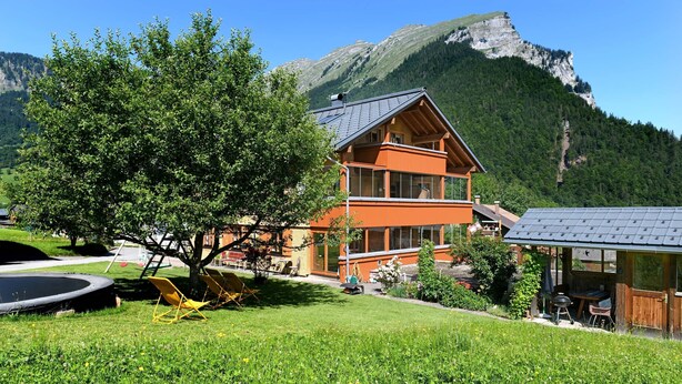Ein Berggasthof mit einem großen Baum im Vordergrund, einem Trampolin und einem Essbereich.