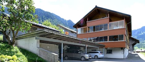 Un edificio de varios pisos con estacionamiento techado, montañas al fondo y un árbol en primer plano.
