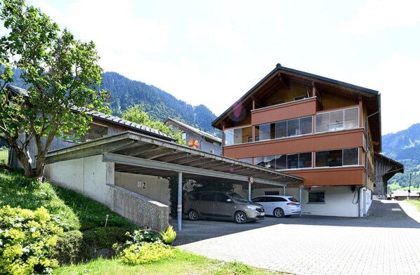 A multi-story building with a covered parking area, a mountain backdrop, and a tree in the foreground.