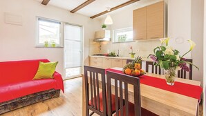 A compact living space with a red sofa, wooden dining table, and chairs. A kitchen area with wooden cabinets and a window with blinds is visible.