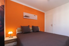 A bedroom with an orange wall, a bed with a brown bedspread, two pillows, a bedside table with a lamp, and a painting above the bed.