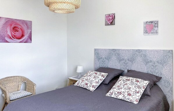 Ein Schlafzimmer mit einem Bett, zwei Kissen, einem Nachttisch, einer Lampe, einem Korbstuhl und drei Wandbildern.