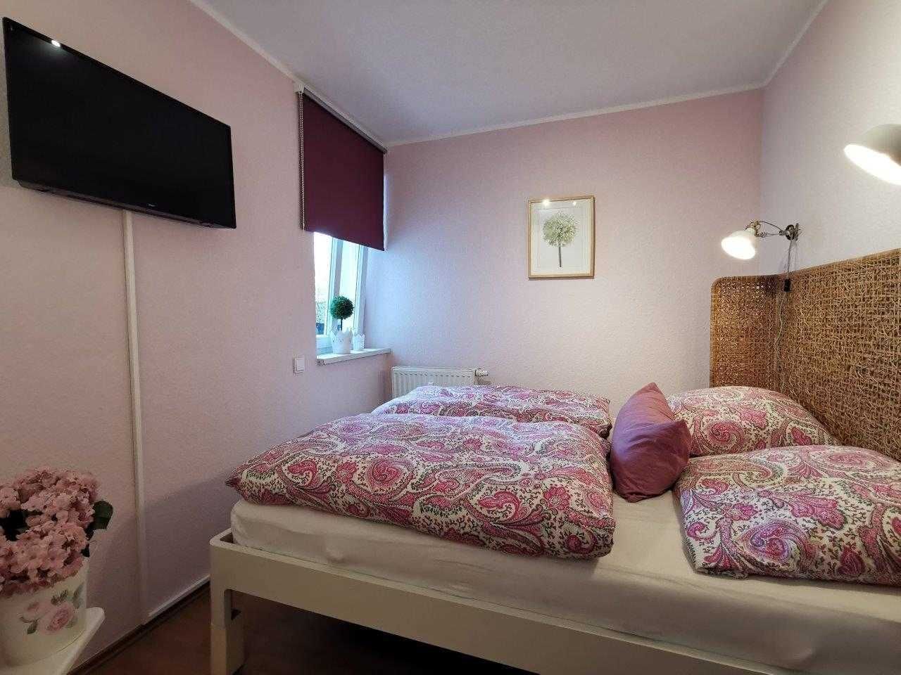A bedroom with a bed, a TV mounted on the wall, a window with a blind, a framed picture on the wall, and a bedside lamp.