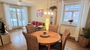 A dining area with a round wooden table and wicker chairs, a sofa in the background, and a window with curtains. - (224) 2-room apartment - apartment house Strandburg (Kühlungsborn)