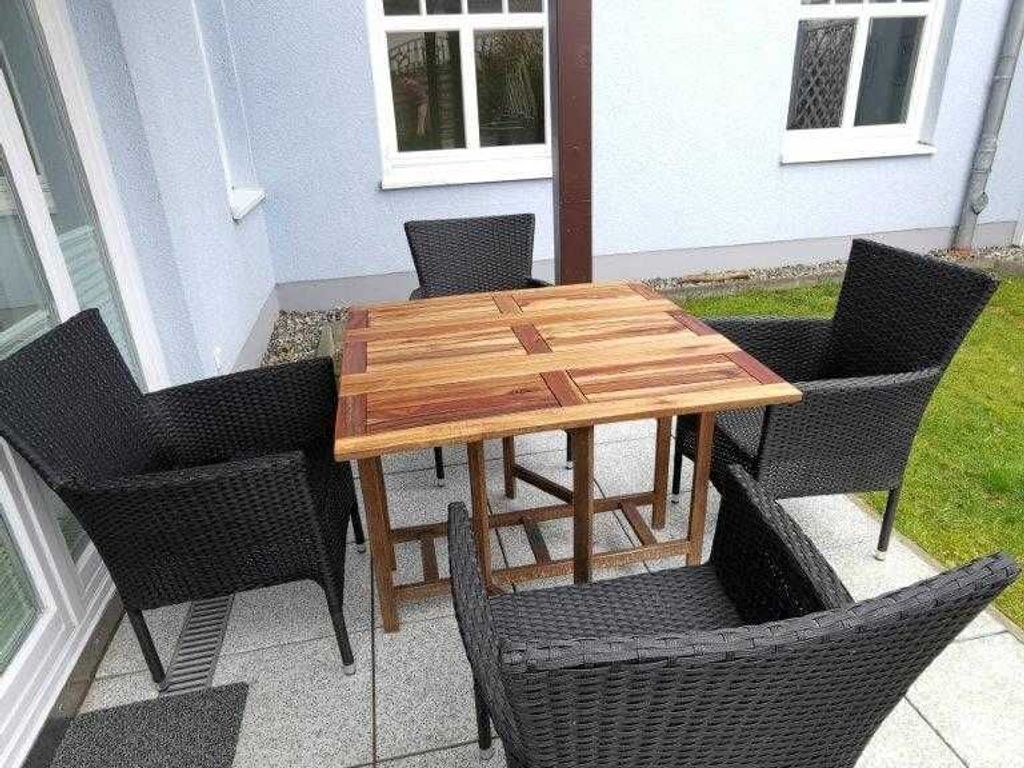 A square wooden table with a woven chair design, surrounded by four chairs, set on a patio with a house in the background.
