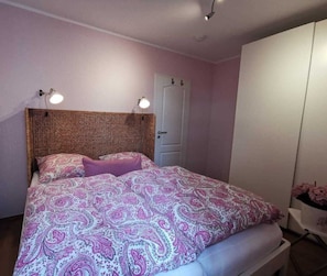 Une chambre à coucher avec une couvre-lit à motifs, une tête de lit en osier, une armoire blanche et une porte dotée d’une poignée.