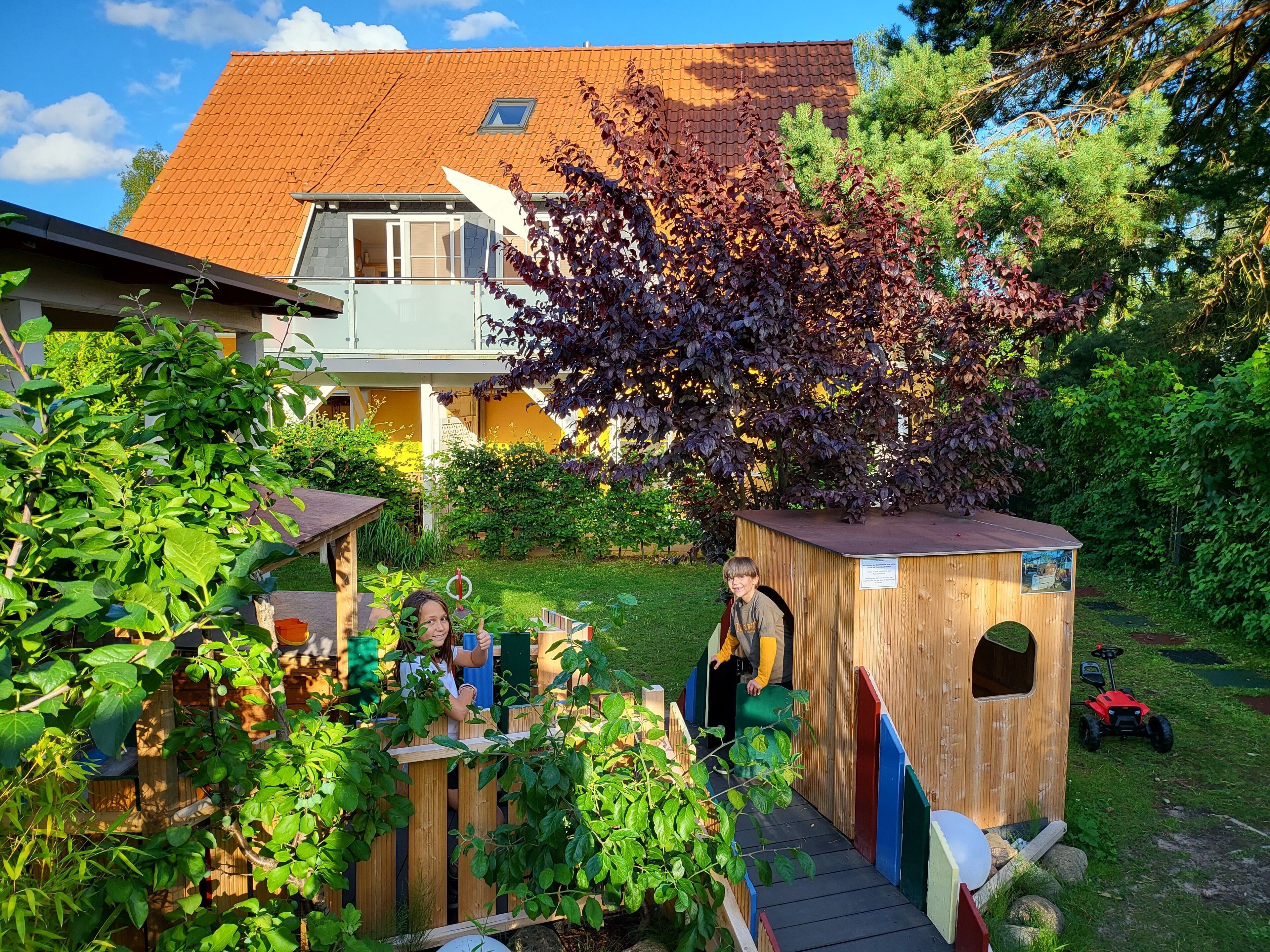 Ein hölzernes Spielhaus mit Rutsche, umgeben von üppigem Grün und einem Haus im Hintergrund.