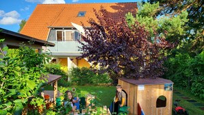 Et legeskjul i træ med rutschebane, omgivet af frodig vegetation og et hus i baggrunden.