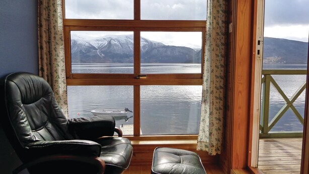 Una habitación con un ventanal grande que da a un lago y montañas, un sillón de cuero y una terraza de madera.