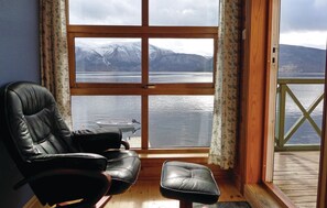 Una habitación con un ventanal grande que da a un lago y montañas, un sillón de cuero y una terraza de madera.
