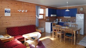 Interior acogedor de una cabaña con un sofá rojo estampado, una mesa de comedor de madera con cuatro sillas, una zona de cocina con armarios azules y un refrigerador blanco, y una ventana pequeña con cortina.