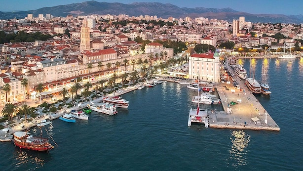 Un paisaje costero con un puerto deportivo, edificios históricos y montañas al fondo.