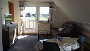 A room with a sofa, a table with a floral tablecloth, a television on a cabinet, and a view of a house through a window.