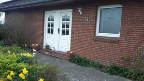A brick house with a white front door, two windows, and a number '12' sign. There are flower pots on the steps and daffodils in the garden.