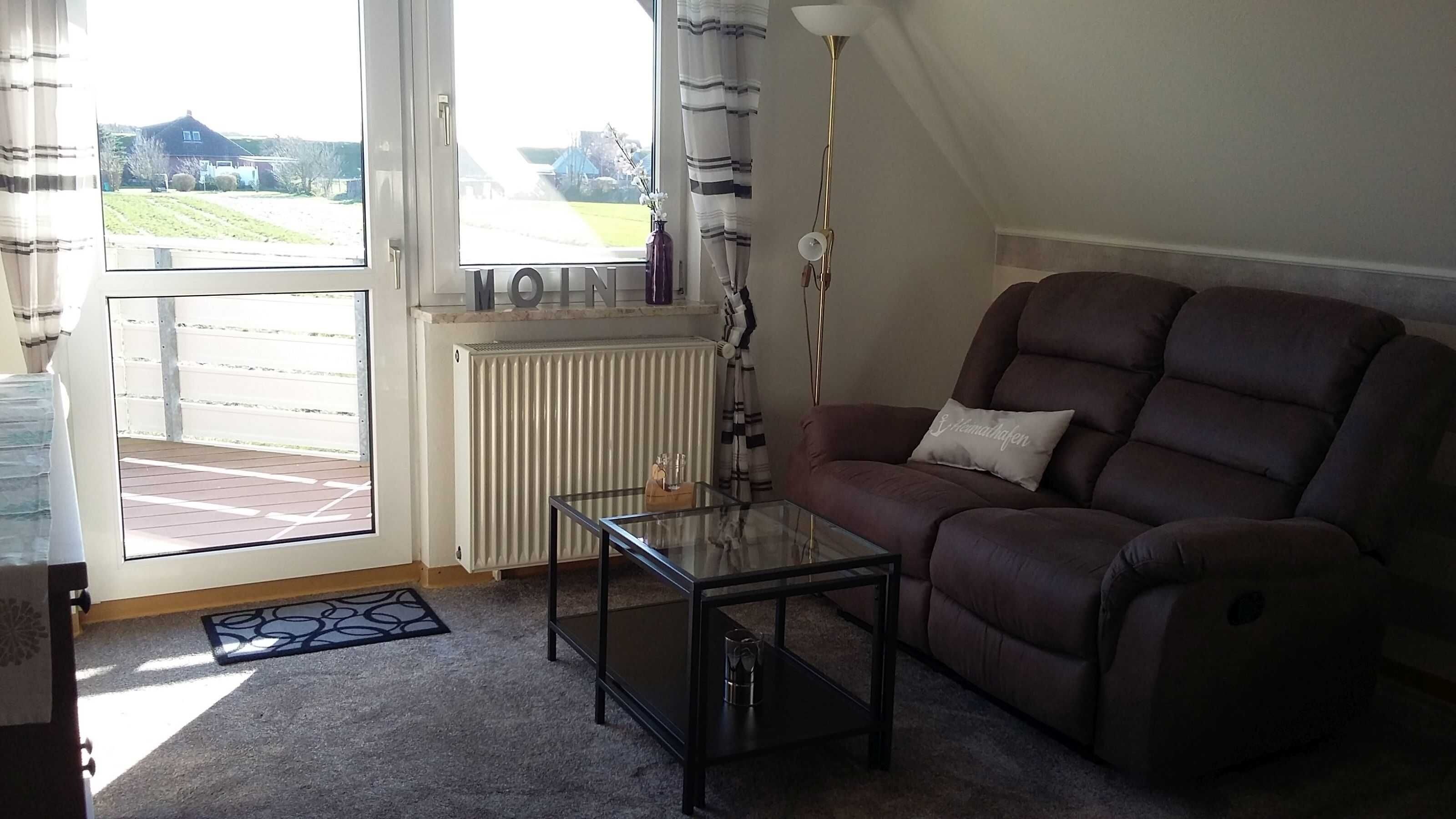 A living room with a brown sofa, a glass coffee table, a radiator, and a view of a house through a window.