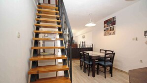 Un escalier avec des marches en bois et des rampes noires mène à l’étage supérieur. On y trouve un coin repas avec une table et des chaises, ainsi qu’un mur orné de tableaux encadrés.