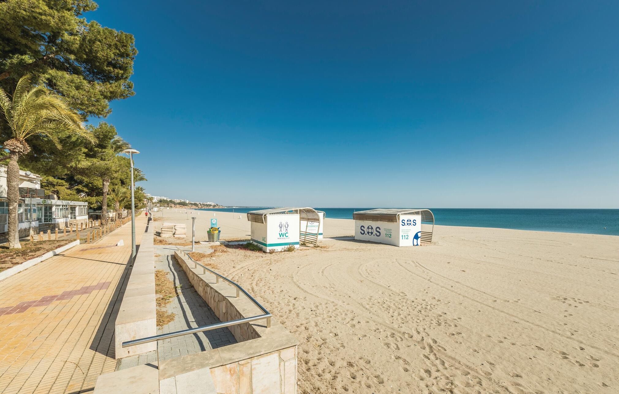 Una spiaggia con un percorso accessibile alle sedie a rotelle che porta a delle cabine designate con le scritte 'WC' e 'SOS'.