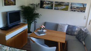 A compact living space with a wooden dining table, a sofa with patterned cushions, a television on a wooden stand, and a potted plant. - Fewo Klein - Holiday home Asel (Vöhl)