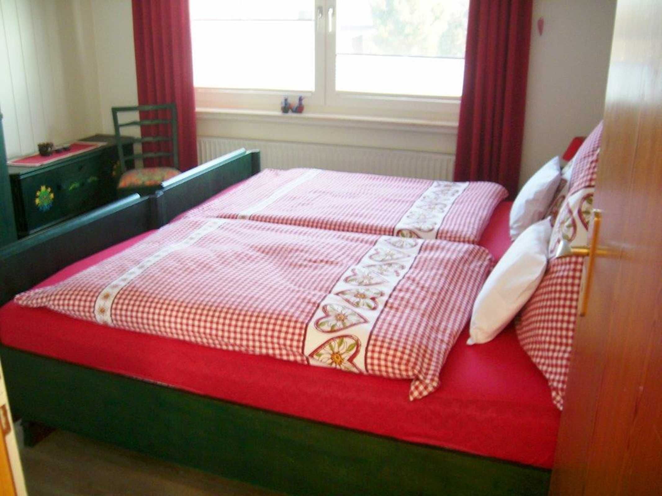 Una cama doble con colcha y almohadas a cuadros rojos y blancos, cabecera de madera y ventana con cortinas rojas.