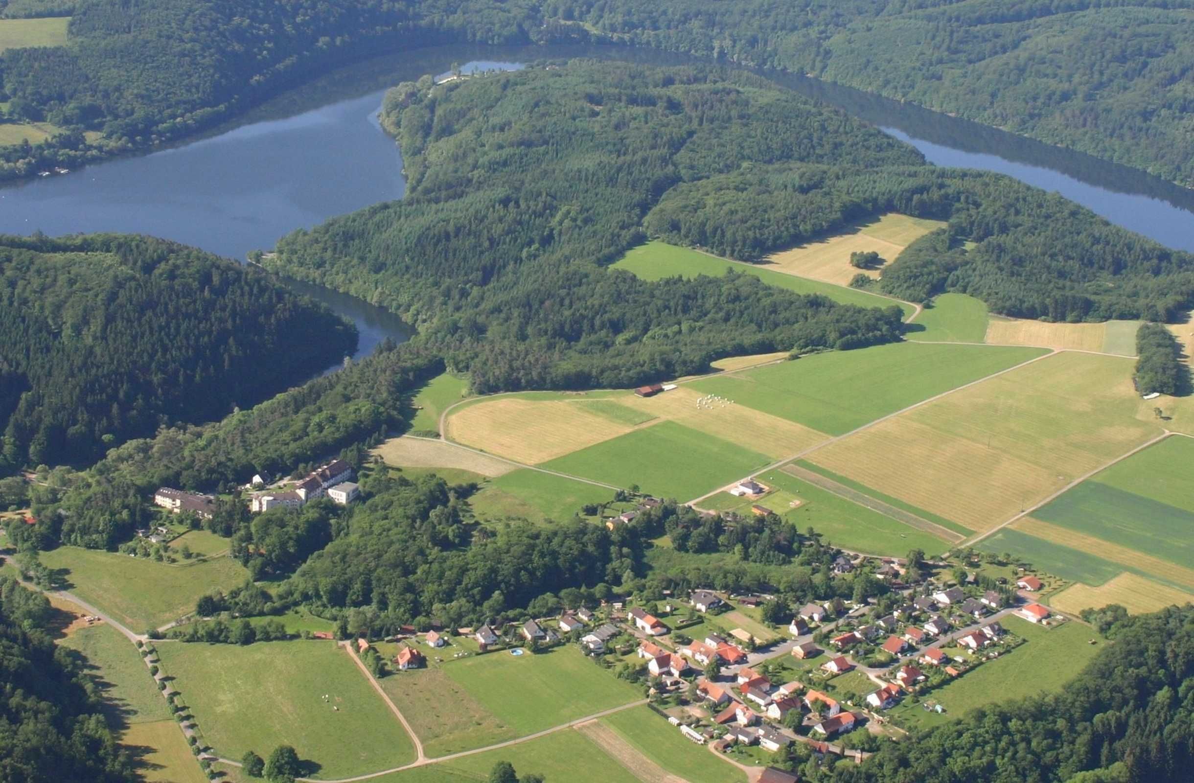 Vue aérienne d’une zone rurale avec une rivière, des champs et un petit village composé de maisons et de bâtiments.