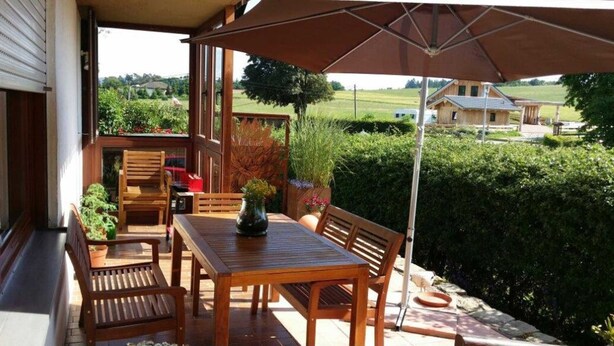 A wooden patio with a table and chairs, a potted plant, and a view of a house and greenery in the background. - Fewo Klein - Holiday home Asel (Vöhl)