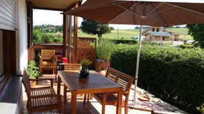 Eine Holzterrasse mit Tisch und Stühlen, einem Blumentopf sowie Blick auf ein Haus und viel Grün im Hintergrund.