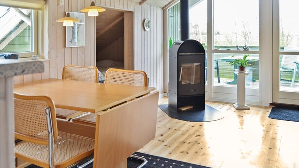 Una habitación acogedora con una mesa y sillas de madera, un estufa de leña empotrada y grandes ventanas que ofrecen vistas al exterior.