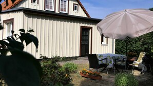 Een wit huis met een rood dak, een overdekte patio met een tafel en stoelen, en een grote parasol.