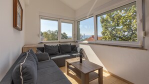 A corner room with a grey sofa, a wooden table with wine glasses, and large windows offering a view of trees and buildings. - Beach front apartment in Njivice with WiFi (Njivice)