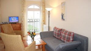 A living room with a sofa, two wicker chairs, a coffee table with a vase of flowers, a television, and a lamp. - (55) 3-room apartment with east-facing balcony - Appartementhaus Atlantik (Kühlungsborn)