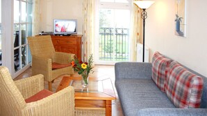 A living room with wicker furniture, a grey sofa, a television, and a view of the outdoors. - (55) 3-room apartment with east-facing balcony - Appartementhaus Atlantik (Kühlungsborn)