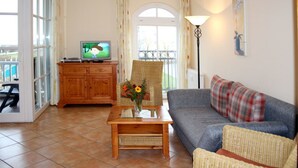 A living room with a sofa, armchair, wooden coffee table, and a television on a cabinet. - (55) 3-room apartment with east-facing balcony - Appartementhaus Atlantik (Kühlungsborn)