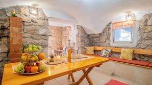 Ein gemütliches Zimmer mit Steinwand, einem Holztisch mit Obst, einer Bank und einem kleinen Fenster mit Vorhängen.