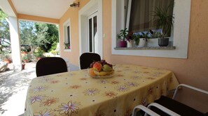 A table with a floral tablecloth, a bowl of fruit, and chairs in a sunny outdoor area with potted plants and a window.