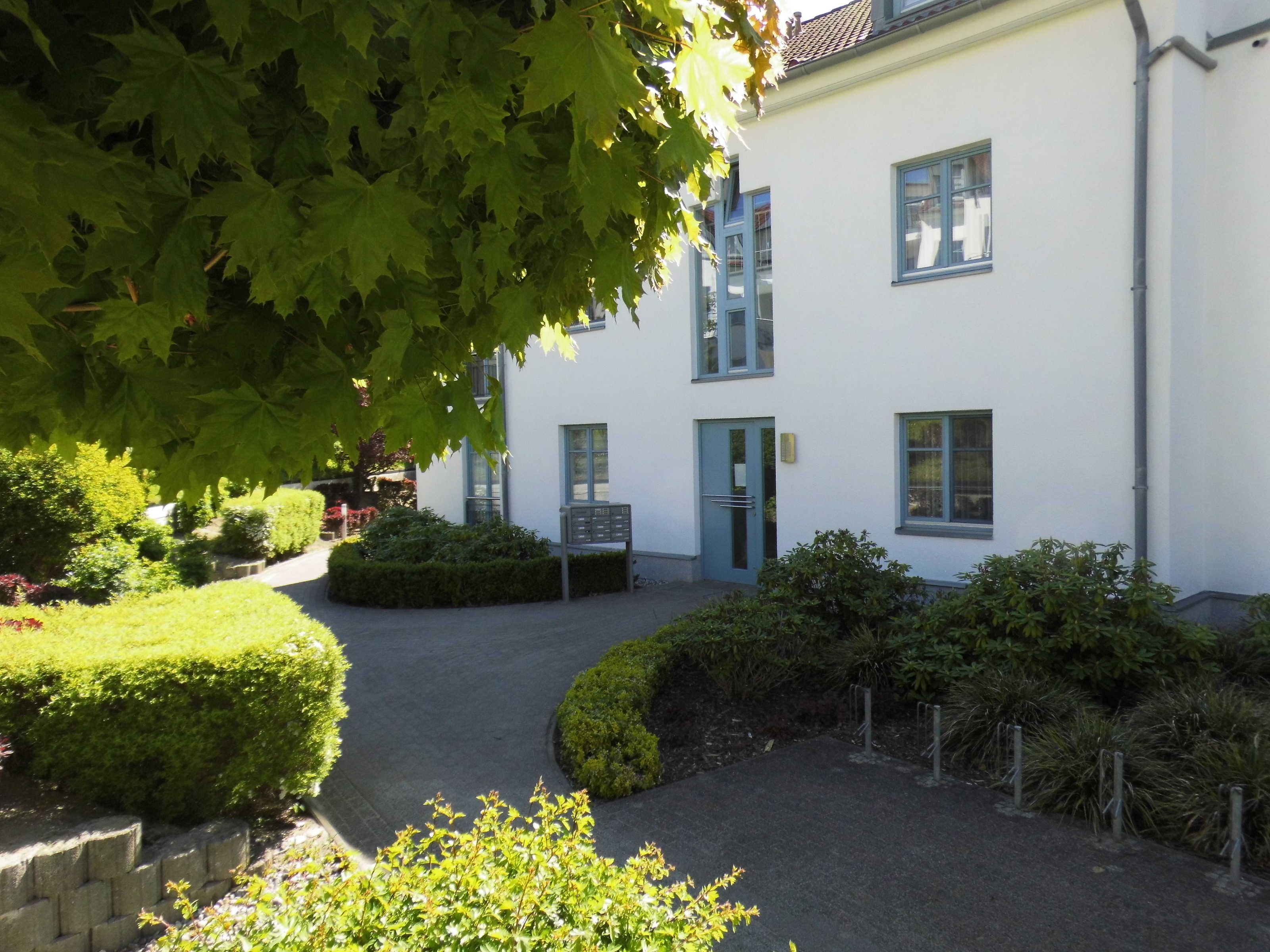 Ein weißes Gebäude mit blauen Fensterrahmen, einem verschlossenen Eingang und einem gepflegten Garten mit Hecken und Bäumen.