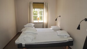 A neatly made bed with white bedding and pillows, a window with curtains, a wall-mounted lamp, and a small table with a lamp. - Lovely home in älmhult with kitchen (Älmhult)