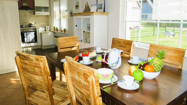 Un coin repas avec une table en bois dressée pour le petit-déjeuner, entourée de chaises en bois. On aperçoit une cuisine à l’arrière-plan, équipée d’appareils électroménagers modernes.