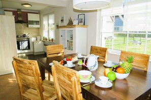 Zona pranzo con tavolo in legno apparecchiato per la colazione, circondato da sedie in legno. Si intravede una cucina sullo sfondo, con elettrodomestici moderni.