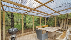 Une terrasse extérieure couverte, avec une table et des chaises en bois, un barbecue et une vue sur une zone boisée.