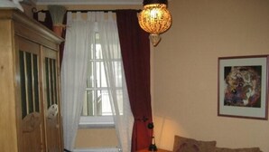 Una camera da letto con un letto, una sedia, un termosifone, una finestra con tende, una lampada e un quadro appeso al muro.