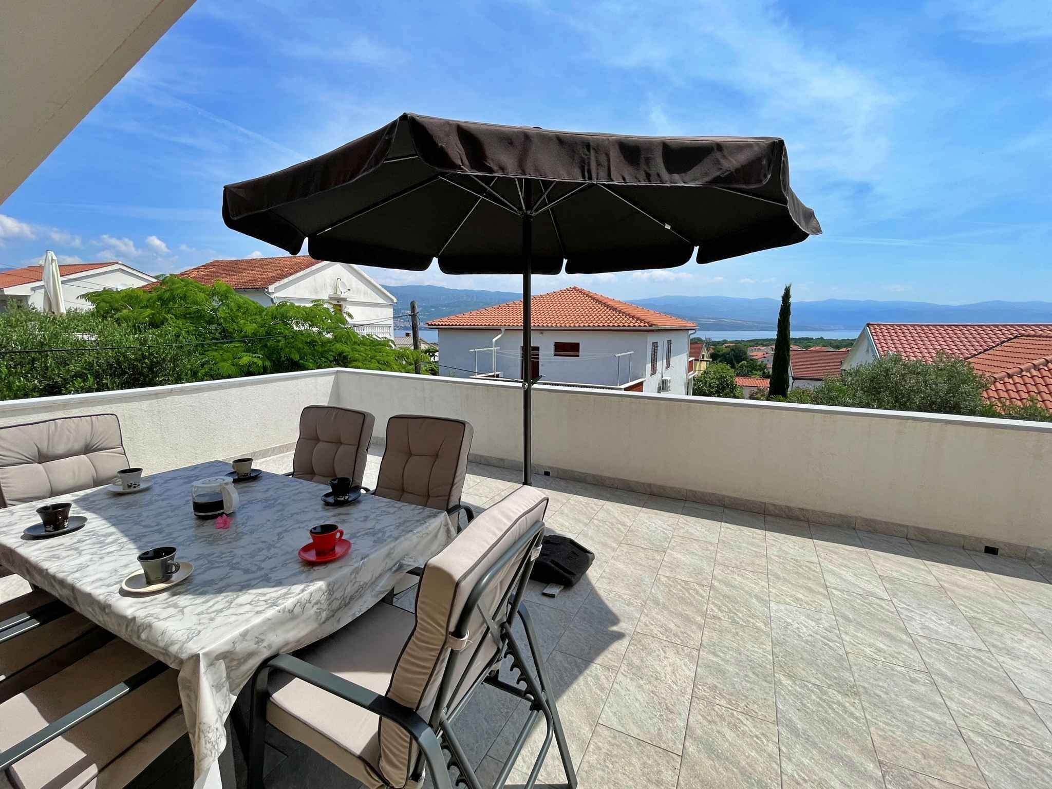 A terrace with a table and chairs, a large umbrella, and a view of houses and mountains.