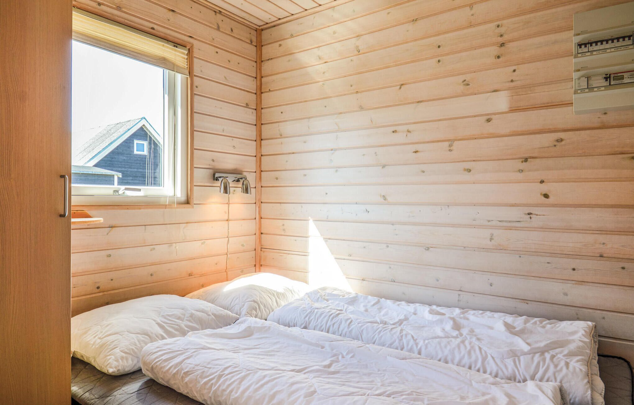 Um quarto pequeno e acolhedor com teto de ripas de madeira, uma cama simples com roupa de cama branca, uma janela com vista para uma casa e um aquecedor fixado na parede.
