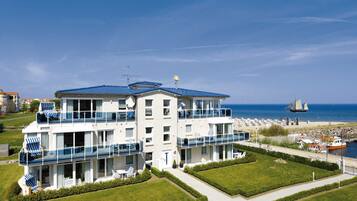 Un hôtel à plusieurs étages doté de balcons, offrant une vue sur la plage et où l’on aperçoit un voilier au loin.