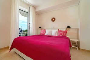 A bedroom with a bed, a nightstand, a lamp, a view through a window, and a decorative wall piece.