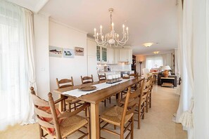 Uma área de refeições com uma longa mesa de madeira, cadeiras, um candelabro e armários de cozinha ao fundo.