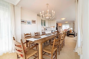 A dining area with a long wooden table, chairs, a chandelier, and kitchen cabinets in the background. - Kein Hüsung (Whg. 1) - Strandresidenz-Kühlungsborn ***** (Kühlungsborn)
