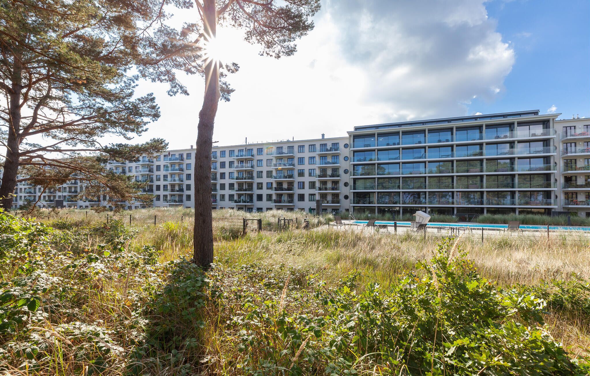 A modern apartment building with multiple balconies, a swimming pool, and a grassy area with trees.