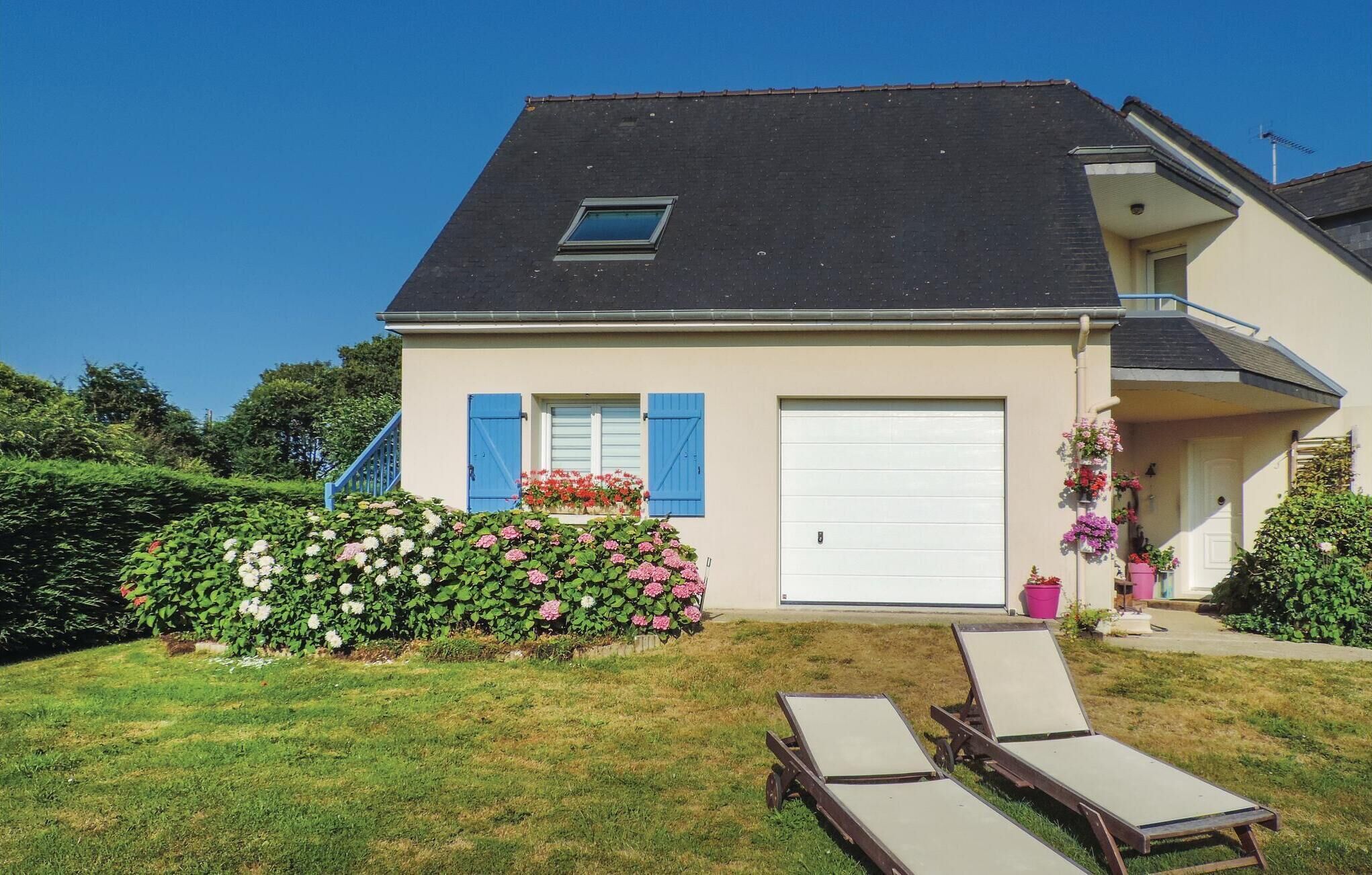 A two-story house with a dark roof, light walls, and a front garage. There is a small window on the upper floor and a blue door on the ground floor. The front yard has a lawn, a flower bed with pink and white flowers, and two lounge chairs.